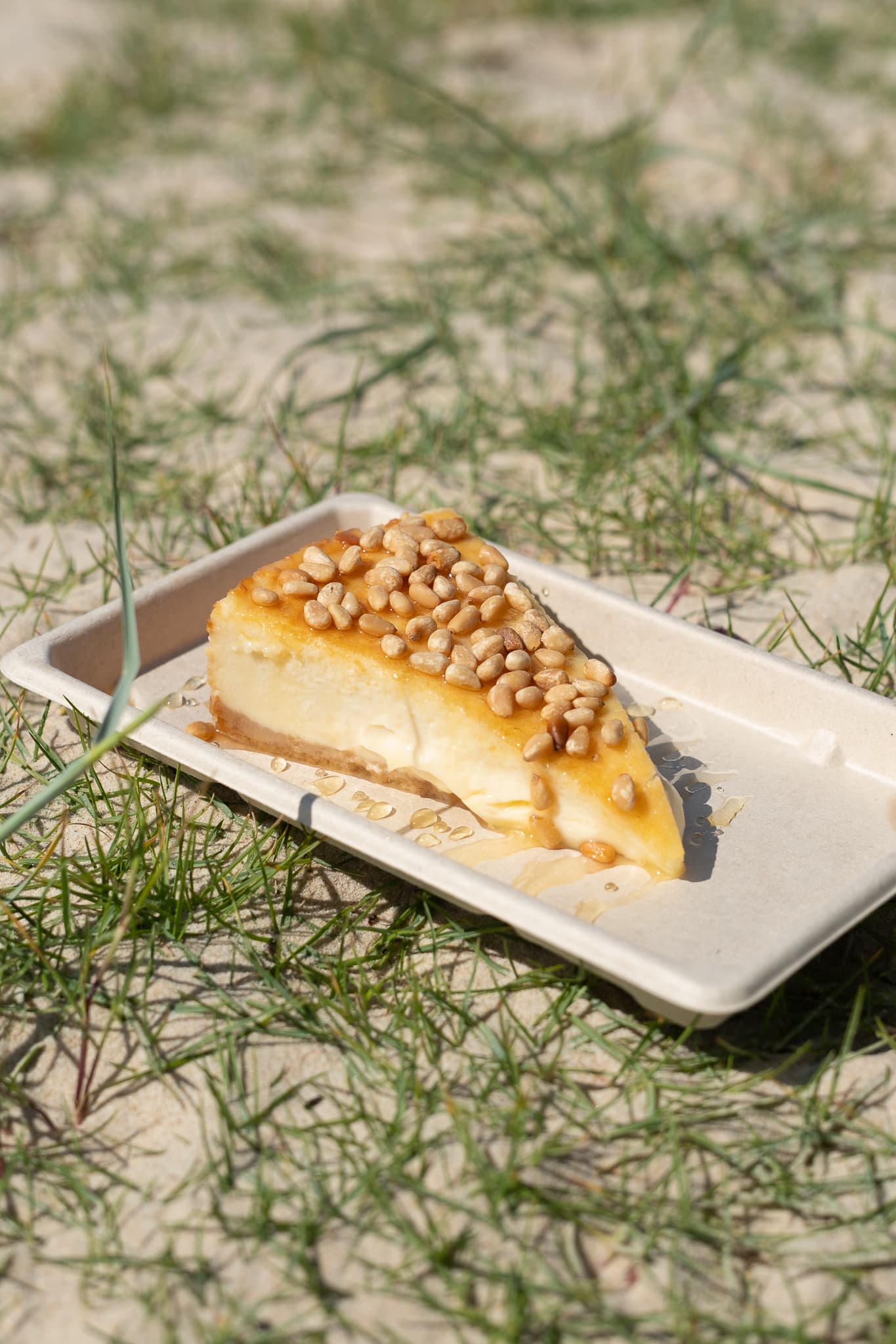Porción de cheesecake con miel y piñones tostados, sobre la arena junto al mar.