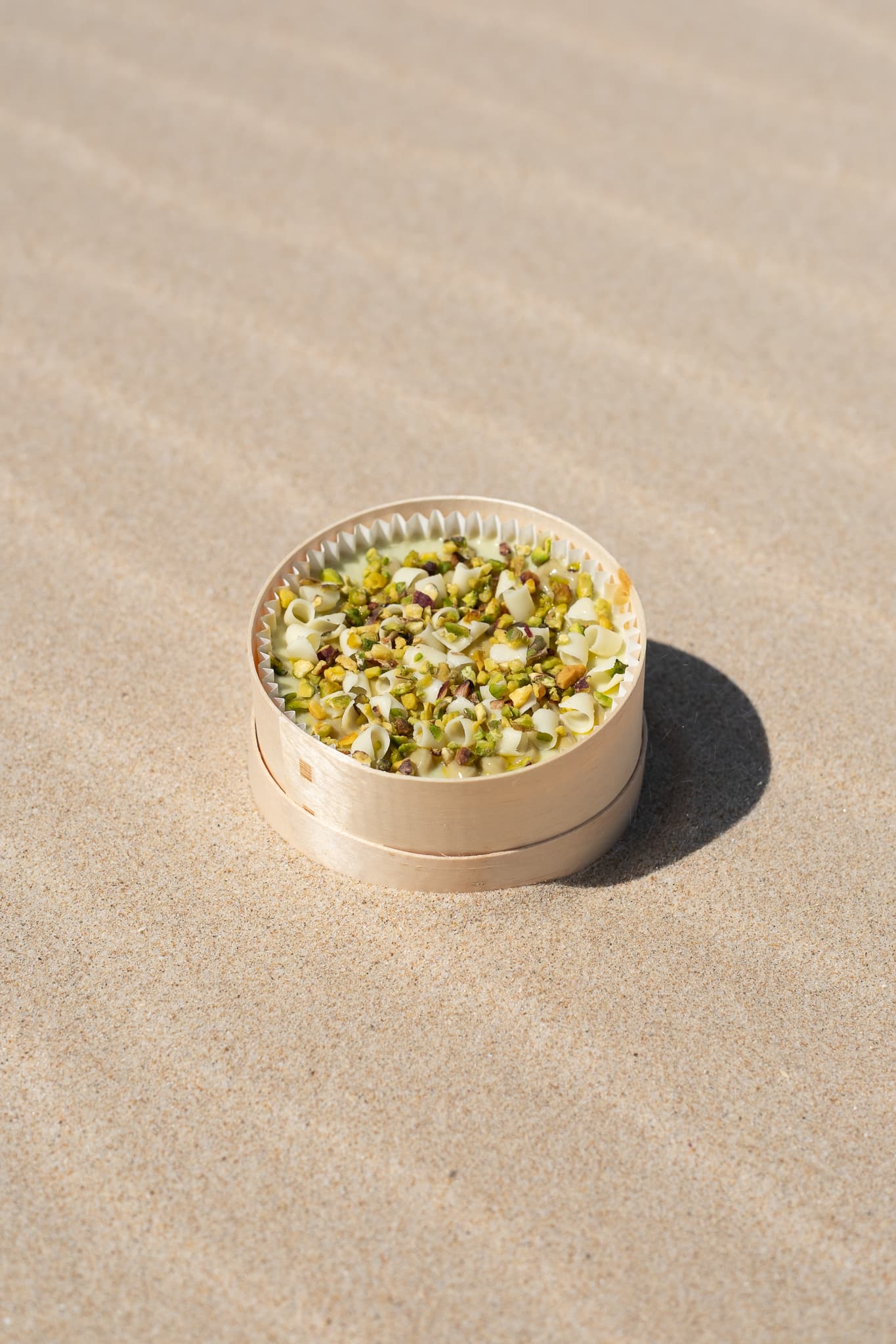 Cheesecake individual cubierto de pistacho troceado, sobre arena de playa.