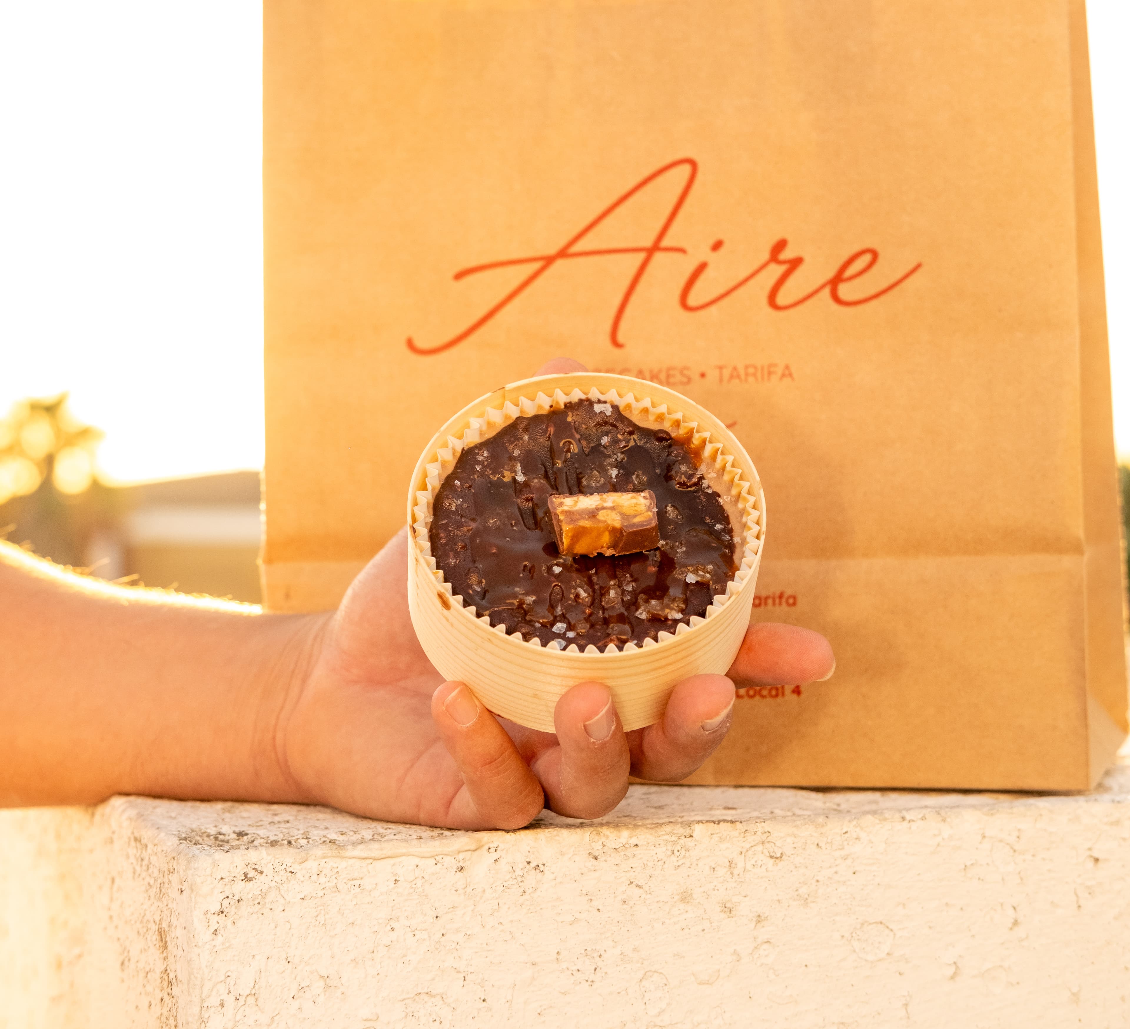 Mano sosteniendo una porción de cheesecake de chocolate y caramelo en su caja de madera, con una bolsa de papel kraft de Aire al fondo, luz cálida de atardecer.
